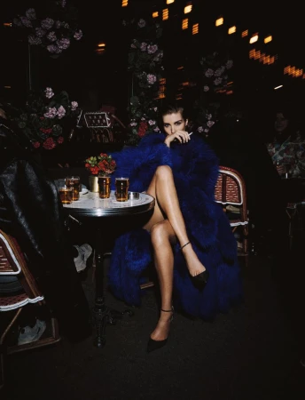 Adult woman with legs crossed sitting at a table with drinks in dim restaurant lighting wearing a blue fur coat and black high heels.