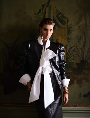 A fashionable woman in a black jacket and white bow stands against a painted backdrop, wearing gold jewelry and stylish earrings.