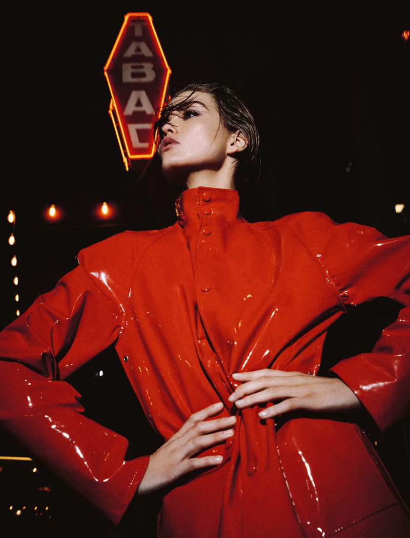 Attractive woman in red latex coat poses at night near a glowing Tabac sign, fashion photography with dark, moody lighting.