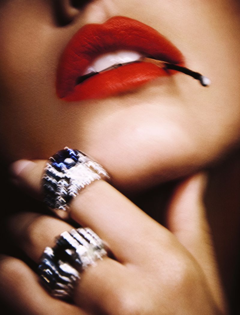 Close-up shot of an adult woman's face with red lipstick, holding a match in her mouth, with rings on her fingers. Beauty shot.