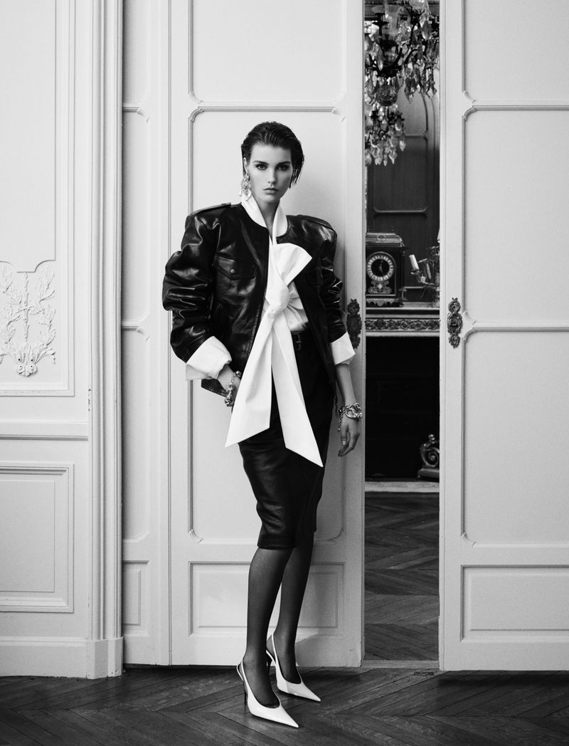 Black and white full shot of a woman in a black jacket with a white bow, skirt, and shoes standing in a doorway indoors.