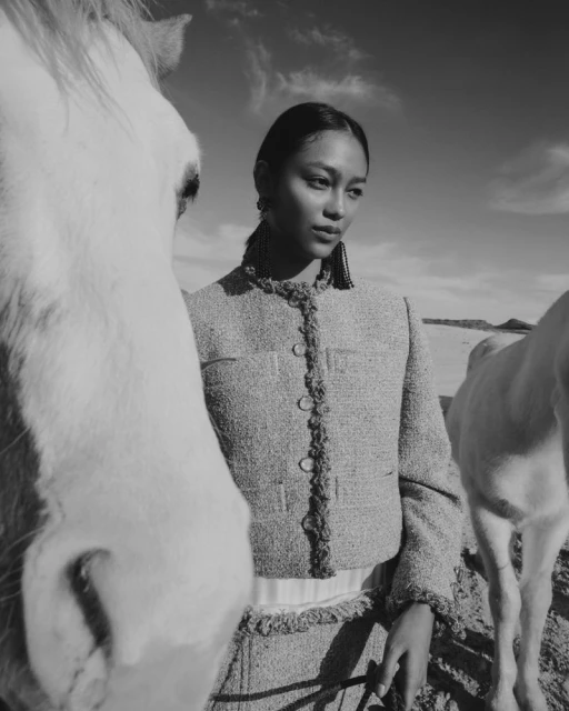 Black and white portrait of a woman in tweed jacket standing next to white horses in a rural landscape, with a clear sky backdrop.