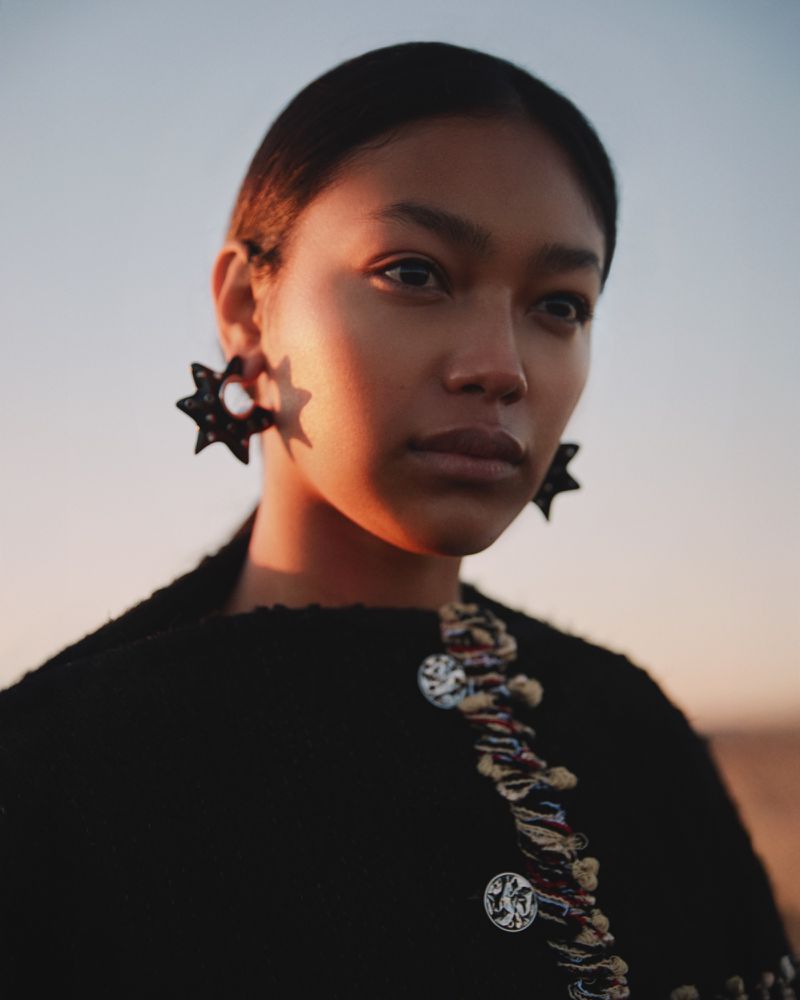 Close-up of a stylish woman wearing star-shaped earrings and a black jacket with ornate buttons against a soft gradient sky at sunset.
