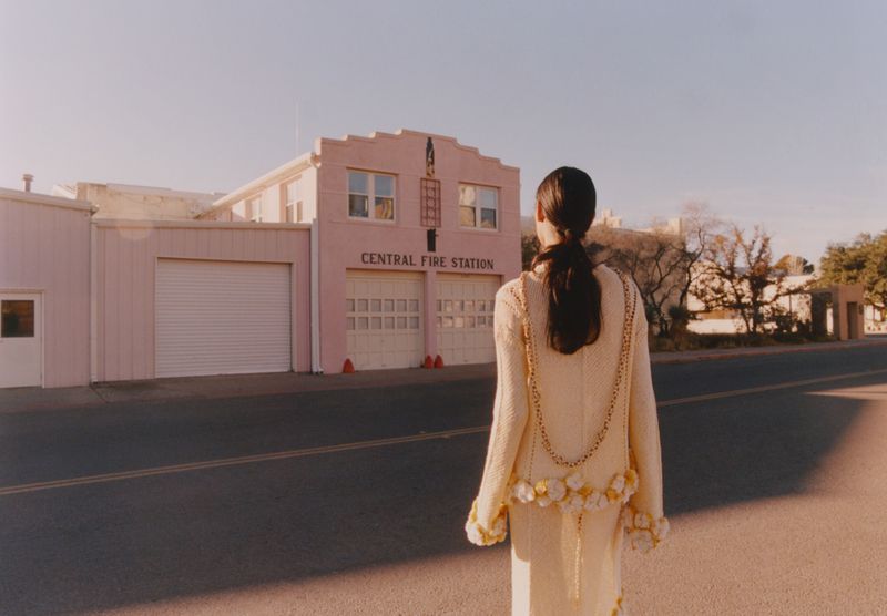 A woman in an elegant cream-colored dress walks down an empty street with a pink Central Fire Station in the background, bathed in golden light.