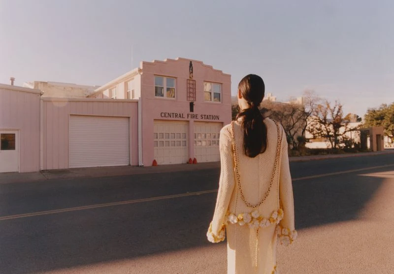 A woman in an elegant cream-colored dress walks down an empty street with a pink Central Fire Station in the background, bathed in golden light.