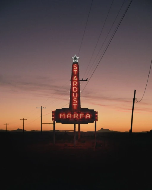 The Stardust Marfa neon sign stands against a dusky sky in a rural landscape, with utility poles and distant mountains visible.