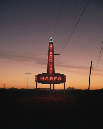 The Stardust Marfa neon sign stands against a dusky sky in a rural landscape, with utility poles and distant mountains visible.