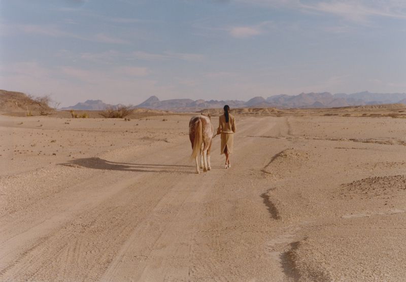 Model walking with a horse on a dusty desert road under an open sky
