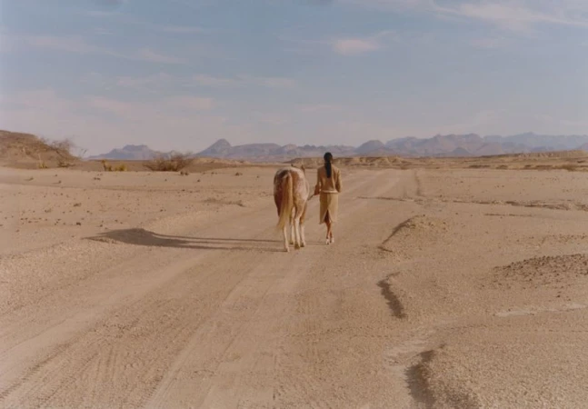 Model walking with a horse on a dusty desert road under an open sky
