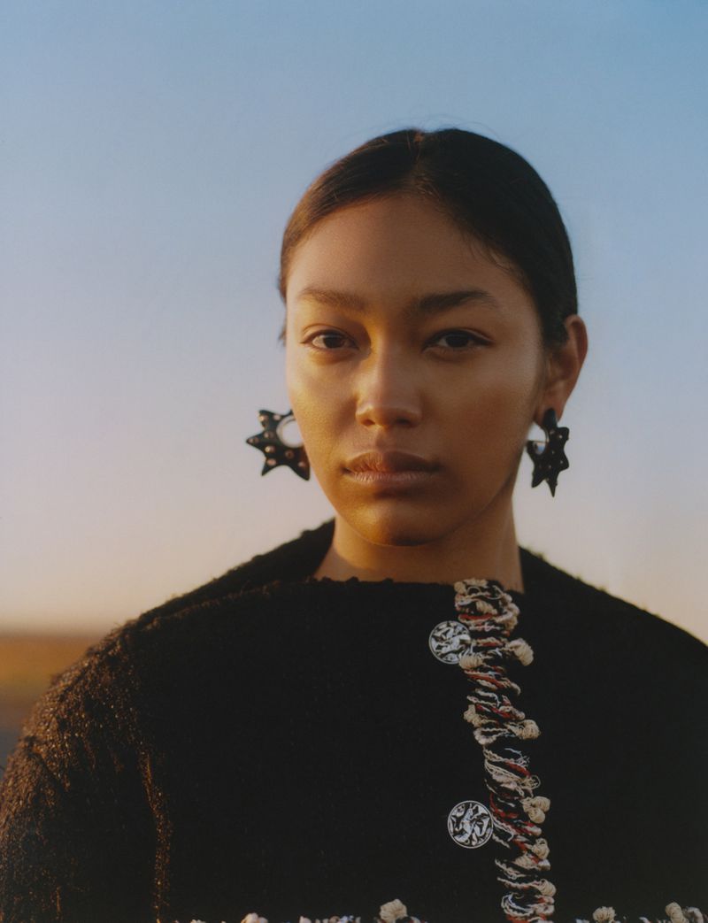 Close-up portrait of a woman with dark hair wearing statement earrings and a black textured jacket in a diffused outdoor light.