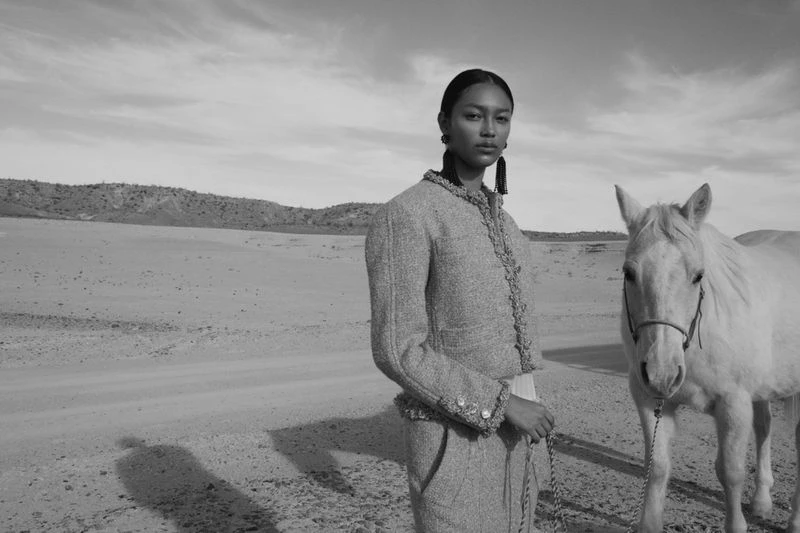 Black and white portrait of an Asian woman in a jacket standing next to a horse in a desert-like landscape underneath a cloudy sky.