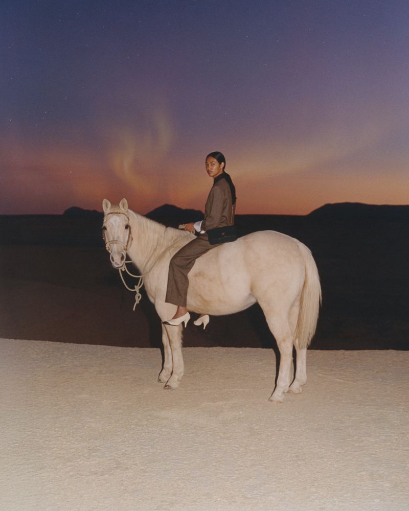 Woman in neutral suit astride pale horse in outdoor setting against a twilight sky with dark hills in the background.