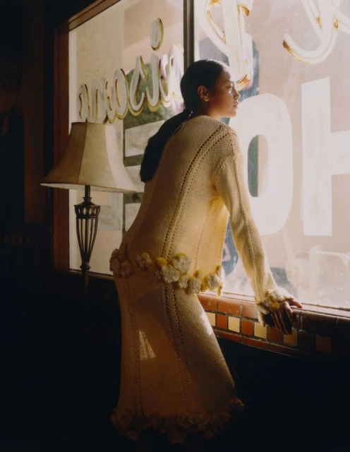 Woman in cream-colored knitwear gazes out window of building, with a lamp and vintage decor casting shadows and warm light.