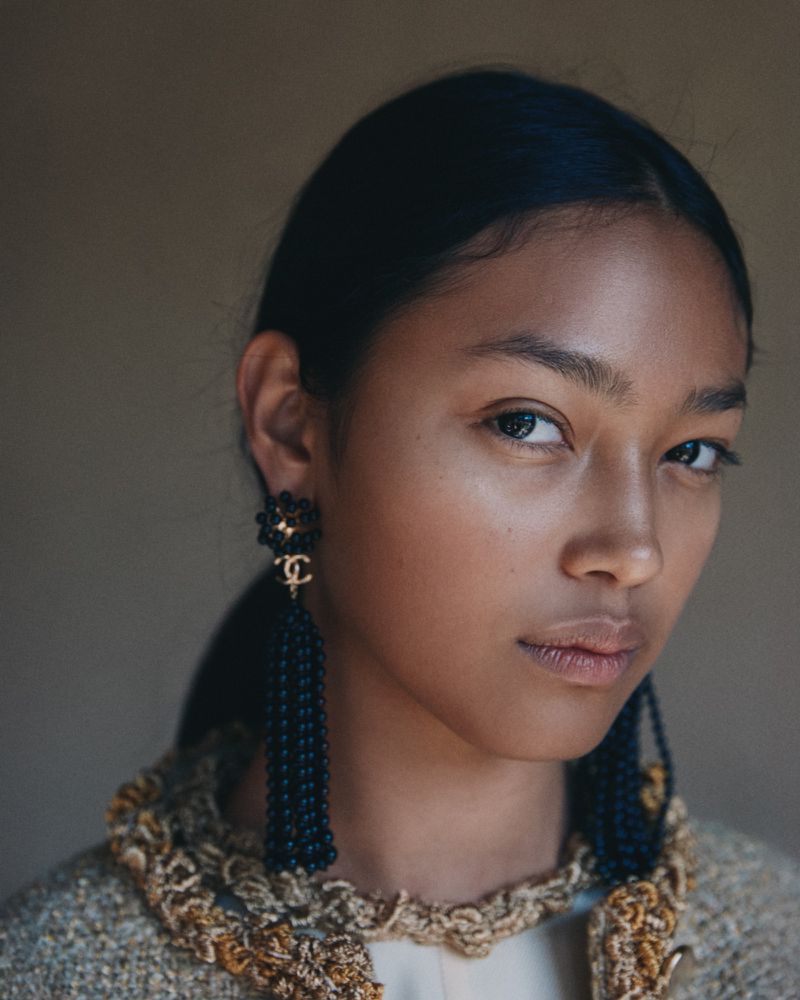 Close-up portrait of a woman with dark hair and light brown skin, wearing dangling earrings and a patterned jacket on a tan backdrop.