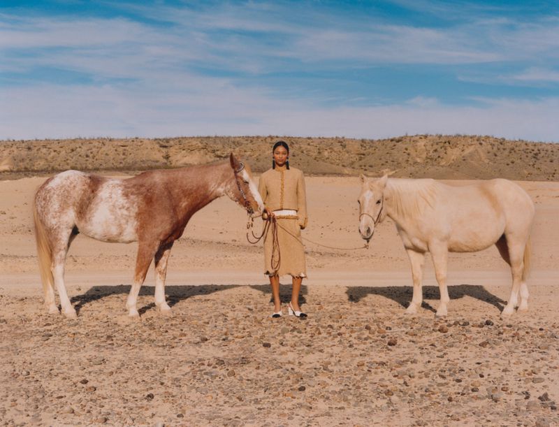 A woman stands between two horses in a desert landscape, wearing a tan skirt suit with the horses in the center of the frame.