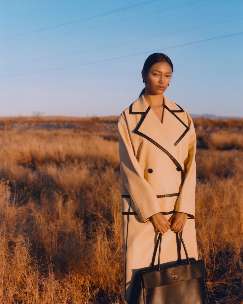 Model wearing a beige coat with black piping and holding a dark handbag in a field under clear blue sky.
