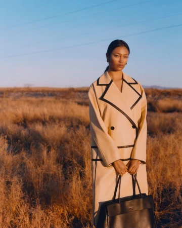 Model wearing a beige coat with black piping and holding a dark handbag in a field under clear blue sky.