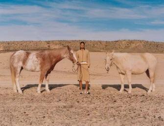 Model wearing a beige tweed skirt suit standing between two horses in an arid landscape with a blue sky.