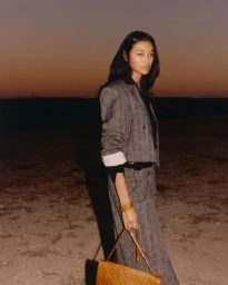 Model standing outdoors at dusk wearing a grey tweed suit and holding a quilted brown shoulder bag with a gold chain strap.