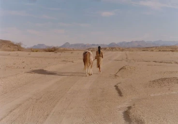 Model walking with a horse on a dusty desert road under an open sky