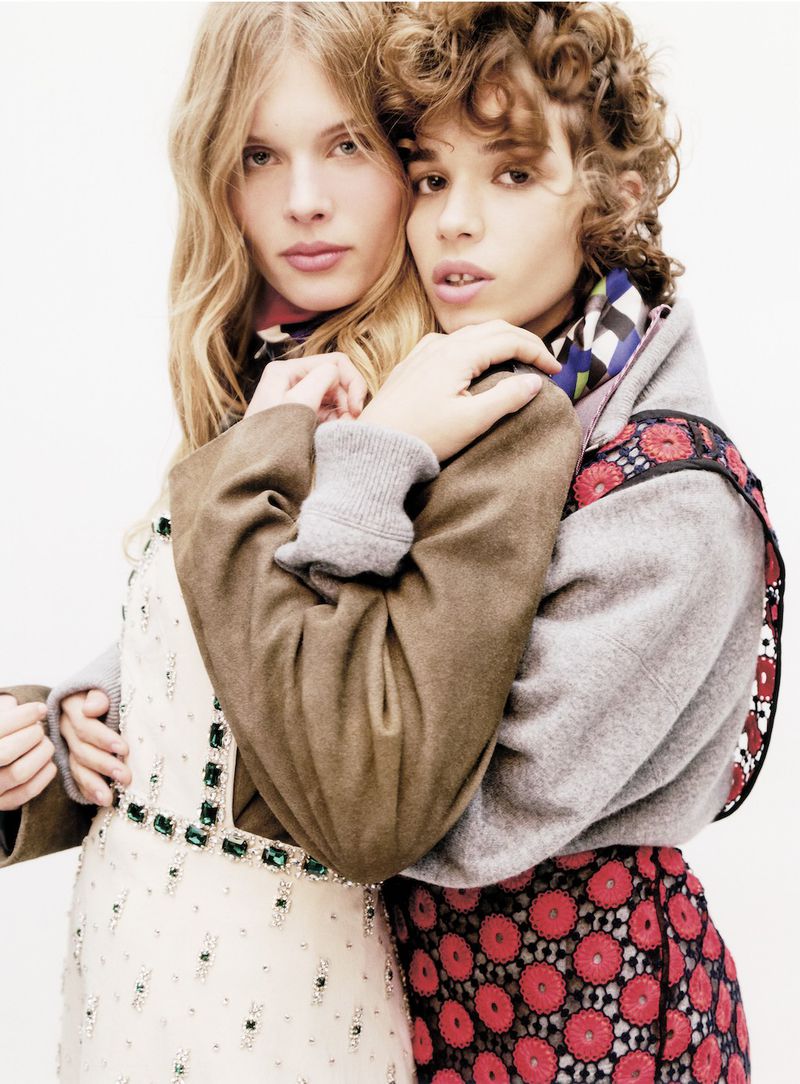 Two fashion models embrace closely in a studio shot, wearing dresses and jackets with intricate details against a neutral backdrop.