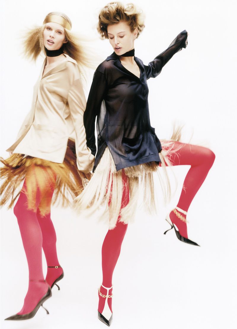 Two women pose in fashionable tops, fringed skirts, tights, and heels against a clean white background in a studio setting.