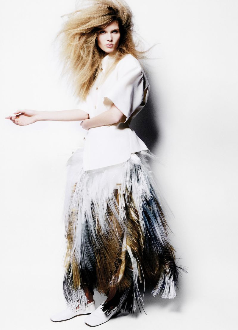 Fashion shot of a female model in a white jacket and skirt with long fringe. She stands in a photo studio against a white backdrop.