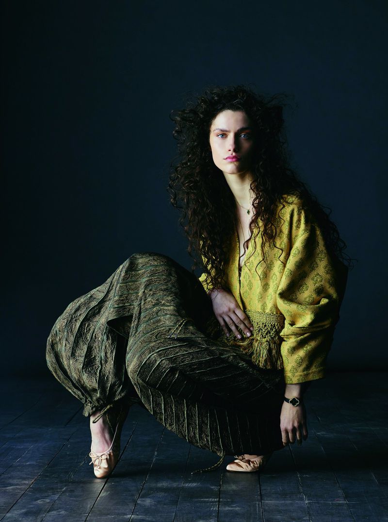 Model with long dark curly hair wearing a mustard patterned top and dark voluminous trousers crouching on a dark floor