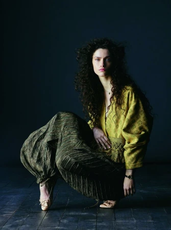 Model with long dark curly hair wearing a mustard patterned top and dark voluminous trousers crouching on a dark floor