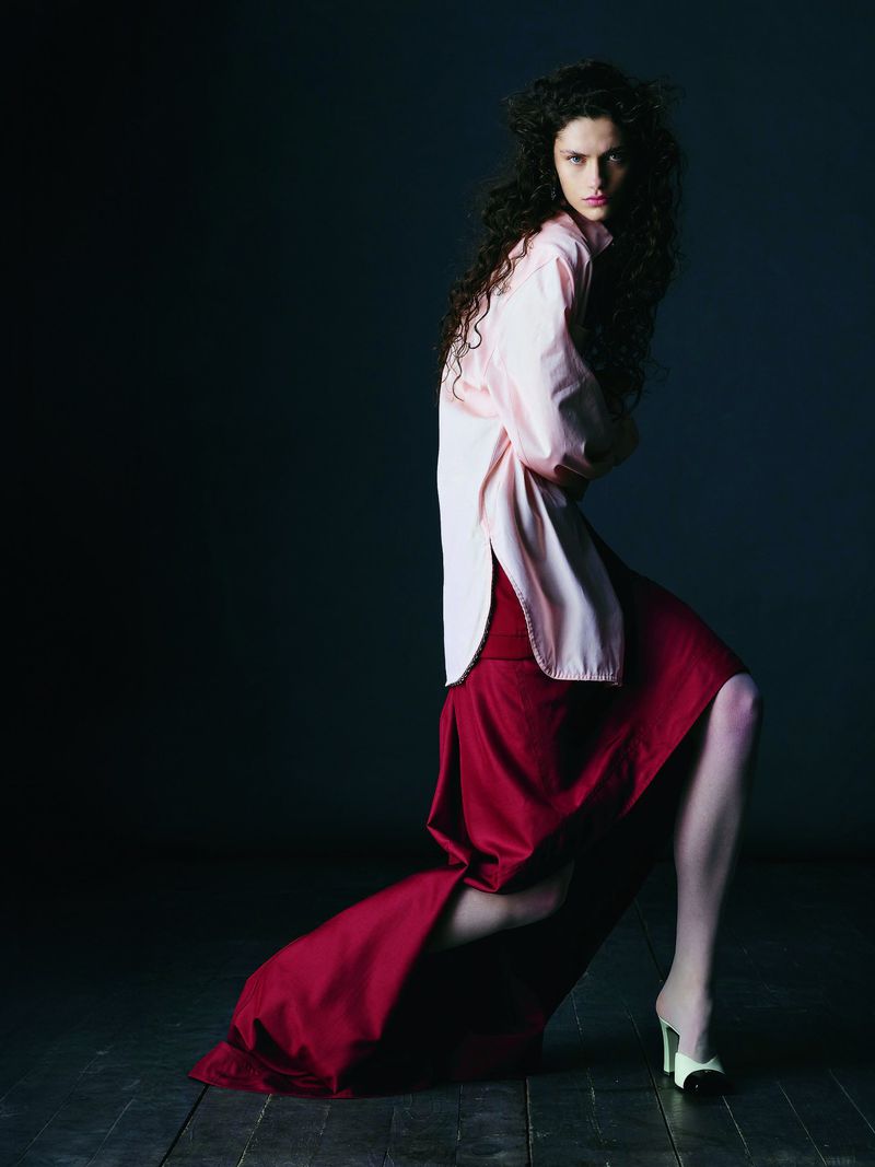 A woman with long dark hair in a pink top and red skirt poses in a dimly lit studio with a dark background on a wood floor.