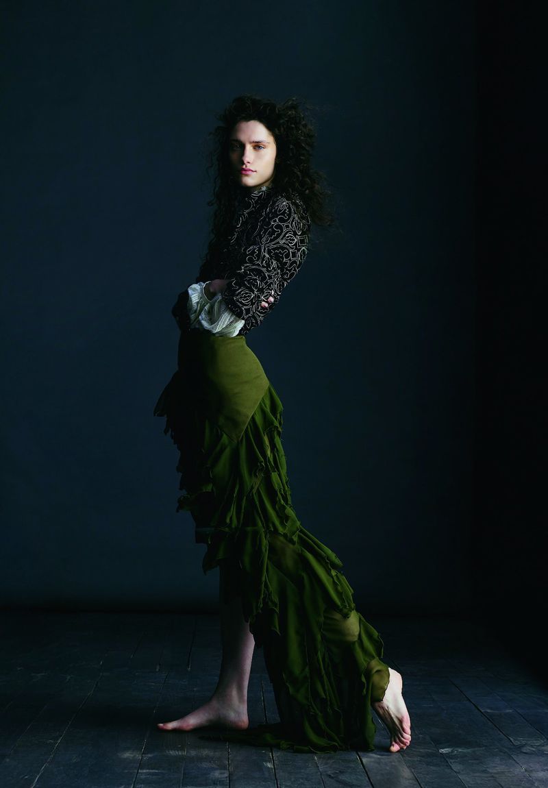 Full-length studio shot of a barefoot human with dark curly hair wearing a black patterned top and long green skirt against dark backdrop.