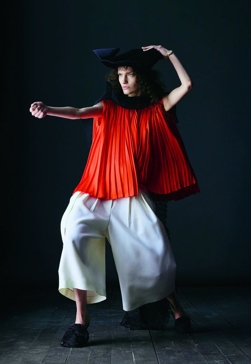 Fashionable individual poses dramatically in a striking red pleated top, white pants, and a black hat against a dark background.