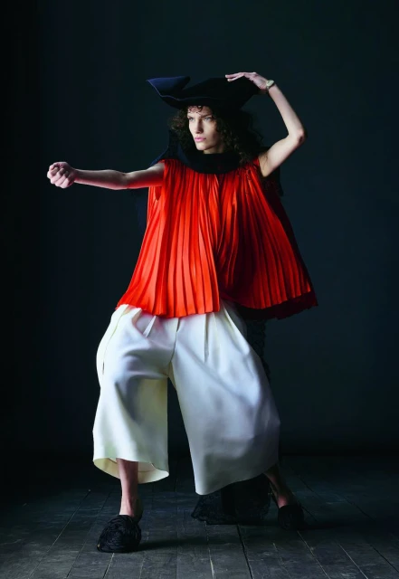 Fashionable individual poses dramatically in a striking red pleated top, white pants, and a black hat against a dark background.
