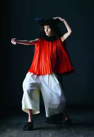 Fashionable individual poses dramatically in a striking red pleated top, white pants, and a black hat against a dark background.