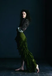 Model with dark curly hair wearing a patterned top and tiered olive green skirt against dark wall and floor