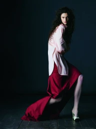Model with long dark curly hair wearing an oversized pink shirt and a long red skirt against a dark background