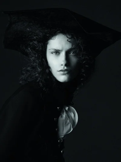 Model with dark curly hair wearing a wide-brimmed hat and dark jacket with ruffles in a dramatic portrait for Telva.