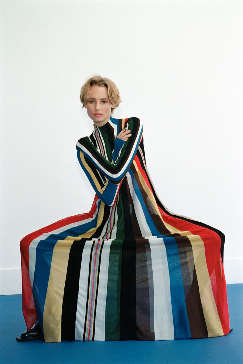 A woman poses in a colorful striped gown with short blonde hair, set against a blue floor and a white wall in a fashion forward display.