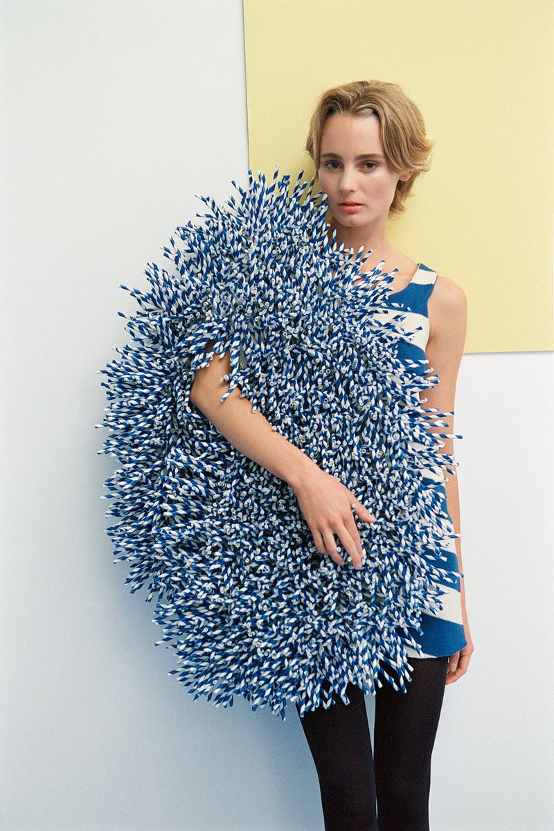 Fashion portrait of a blonde woman wearing a patterned dress, holding a large round object with blue and white spikes against a white wall.