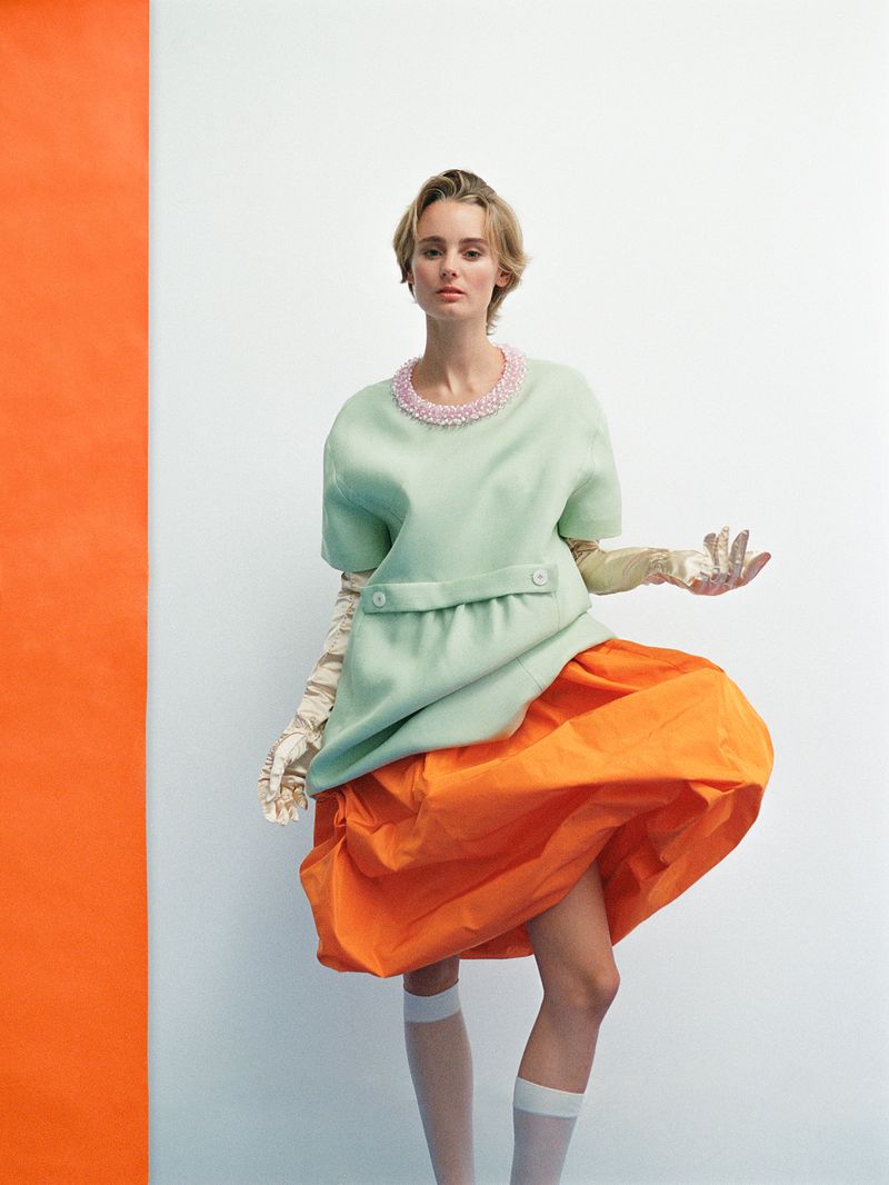 Fashion model in pastel green top with pink beaded collar and orange skirt in a studio setting with a white and orange background.