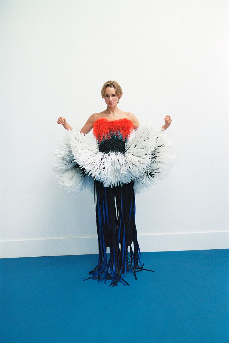 Woman in avant-garde dress with red, black, and white feathery textures poses against a white and blue studio backdrop with a blank expression.