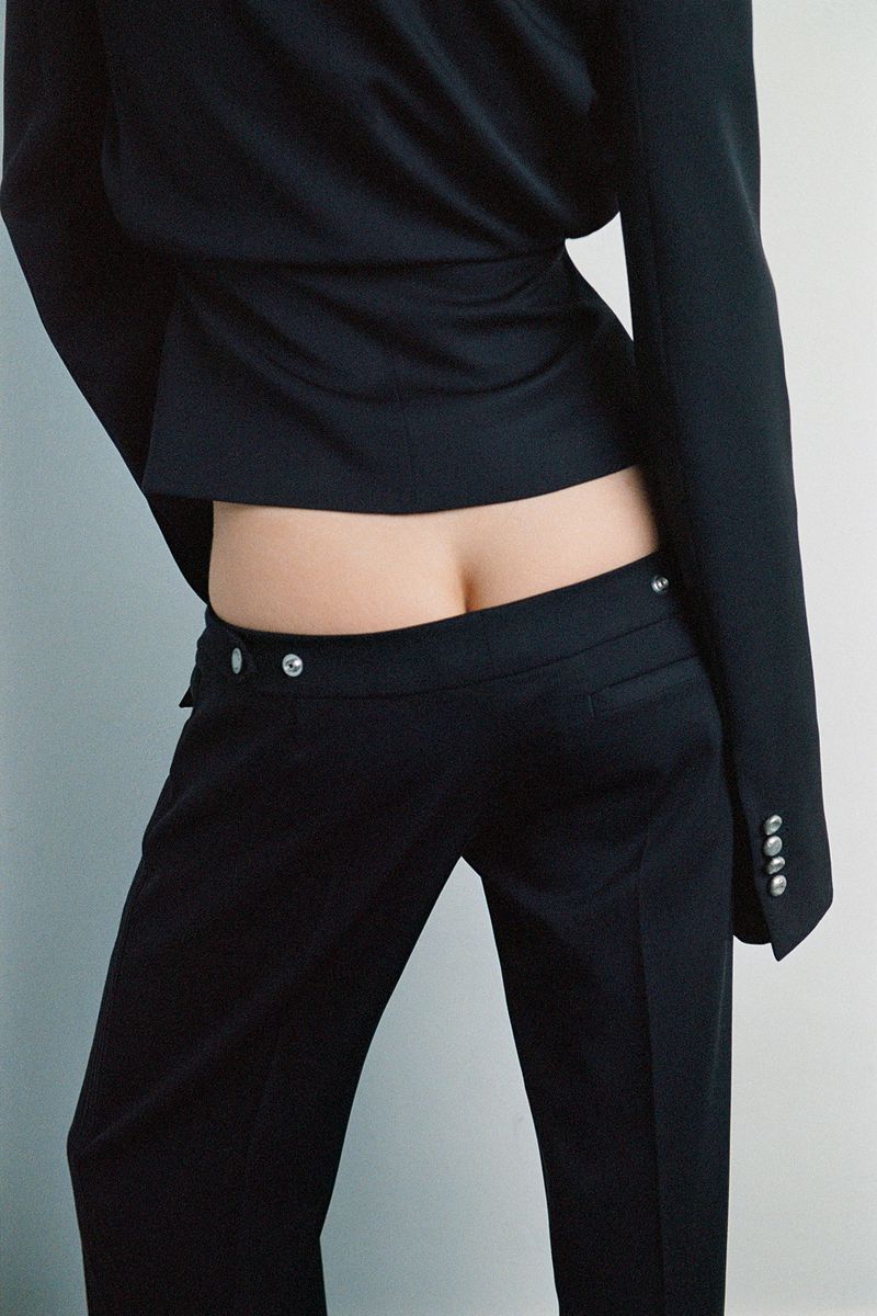 Fashion shot of a woman wearing a black suit with low-rise pants, showing her lower back. The shot is on a simple background.