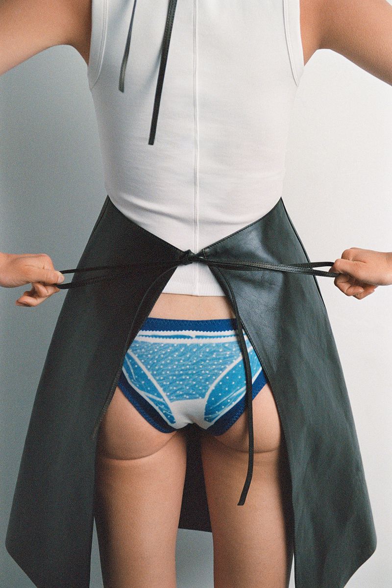 Back view of an unrecognizable person in a white tank top and blue patterned underwear, wearing a black apron tied at the waist.