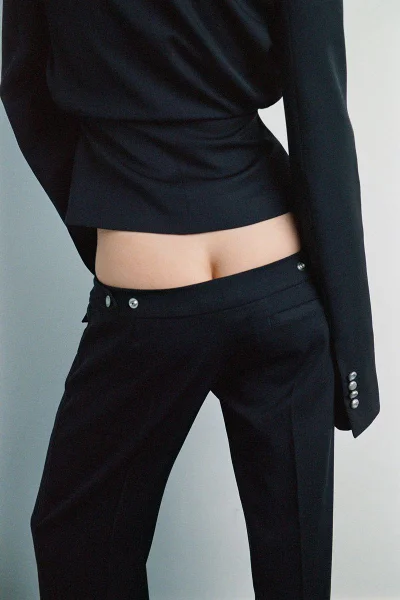 Close-up rear view of a person wearing low-rise black trousers and a black jacket against a neutral grey studio background.