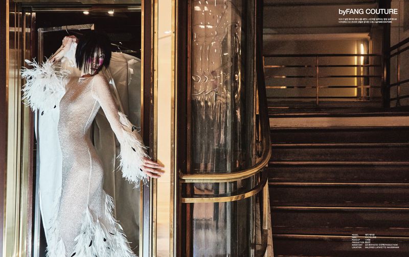 A woman in a feather-trimmed gown stands near a glass elevator in a dimly lit, elegant building with a staircase in the background.