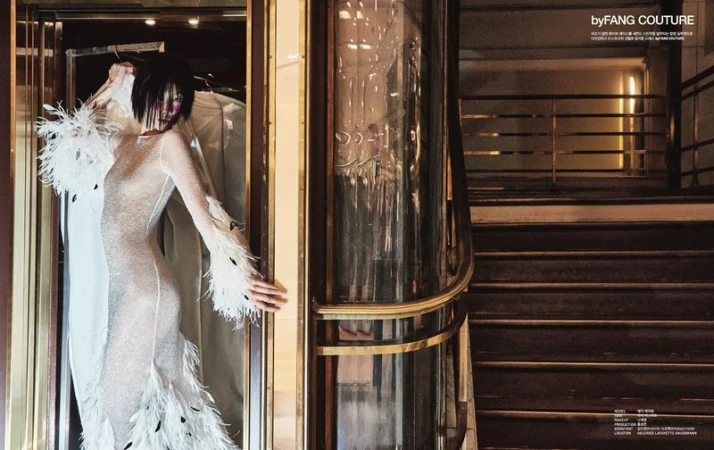 A woman in a feather-trimmed gown stands near a glass elevator in a dimly lit, elegant building with a staircase in the background.