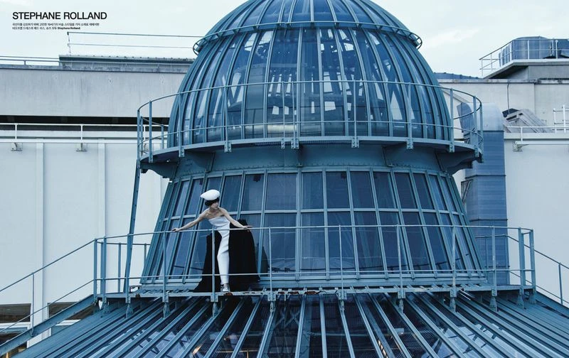 A woman in a dress and hat poses on a glass-domed building, captured in an architectural fashion shot with bright light and sky.