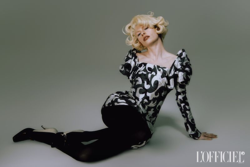 Elegant adult female fashion model poses in a silver and black patterned dress and black tights on a grey studio background.
