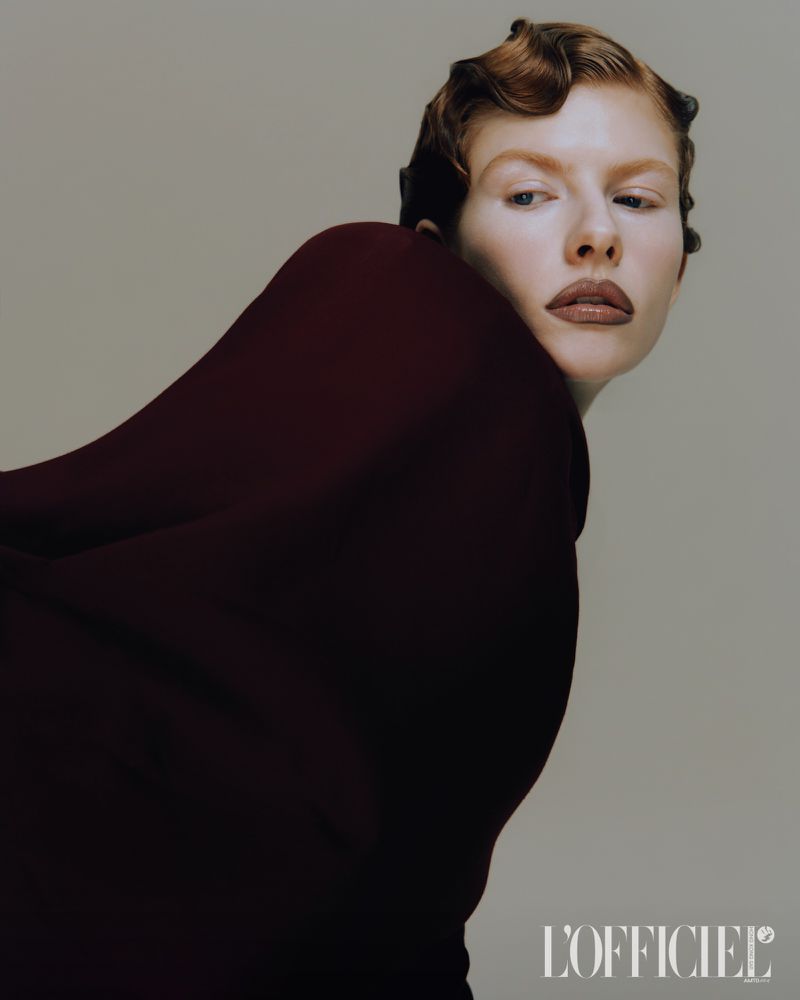 Fashion portrait of a red-haired woman with dark red clothing against a neutral background, posing for a beauty-themed studio shot.
