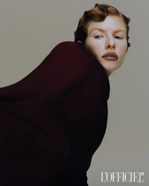 Fashion portrait of a red-haired woman with dark red clothing against a neutral background, posing for a beauty-themed studio shot.